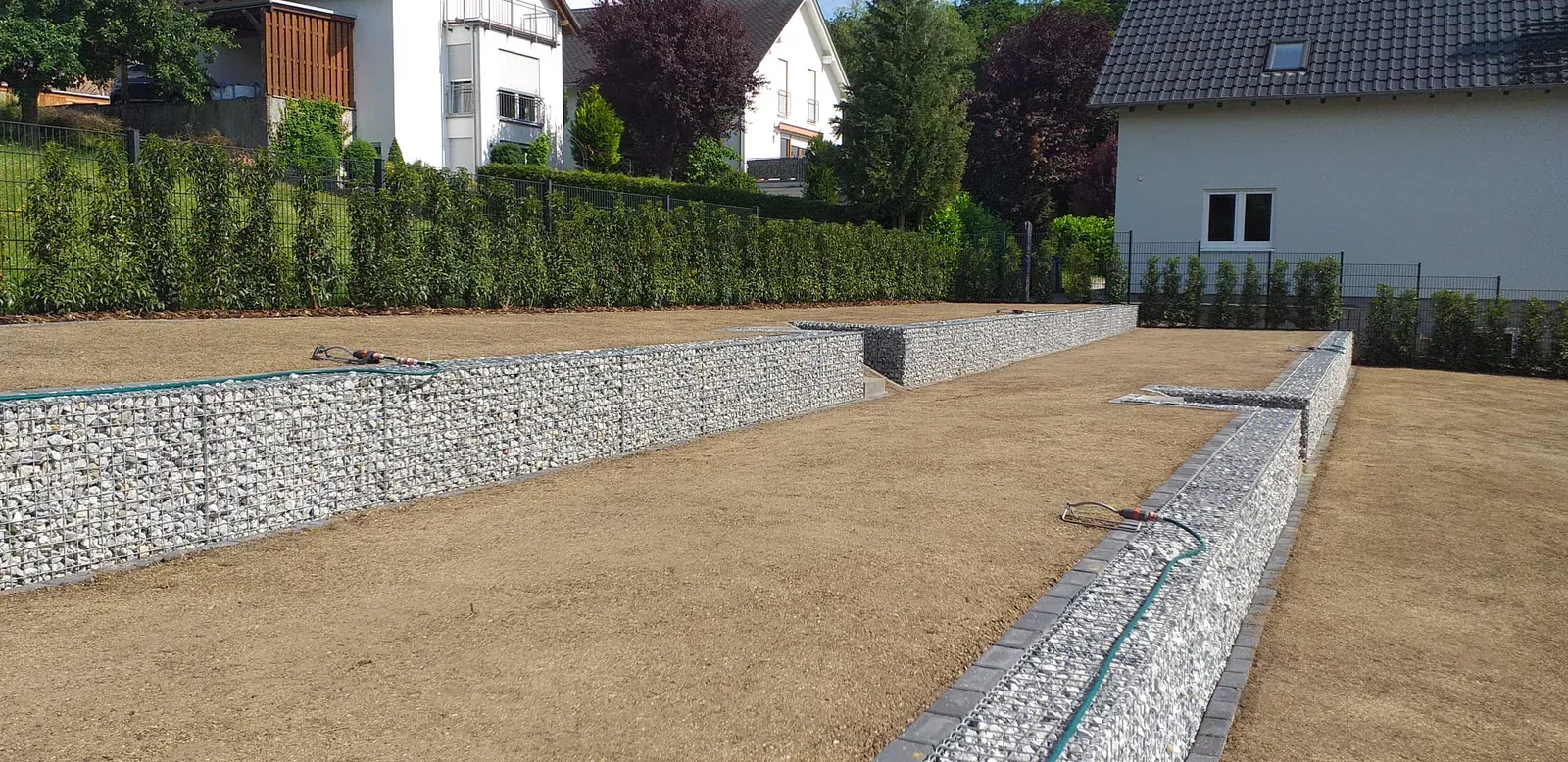 Fertige Gabionenabfangung vor einer eben vorbereiteten Gartenfläche und bestehender Heckenstruktur.