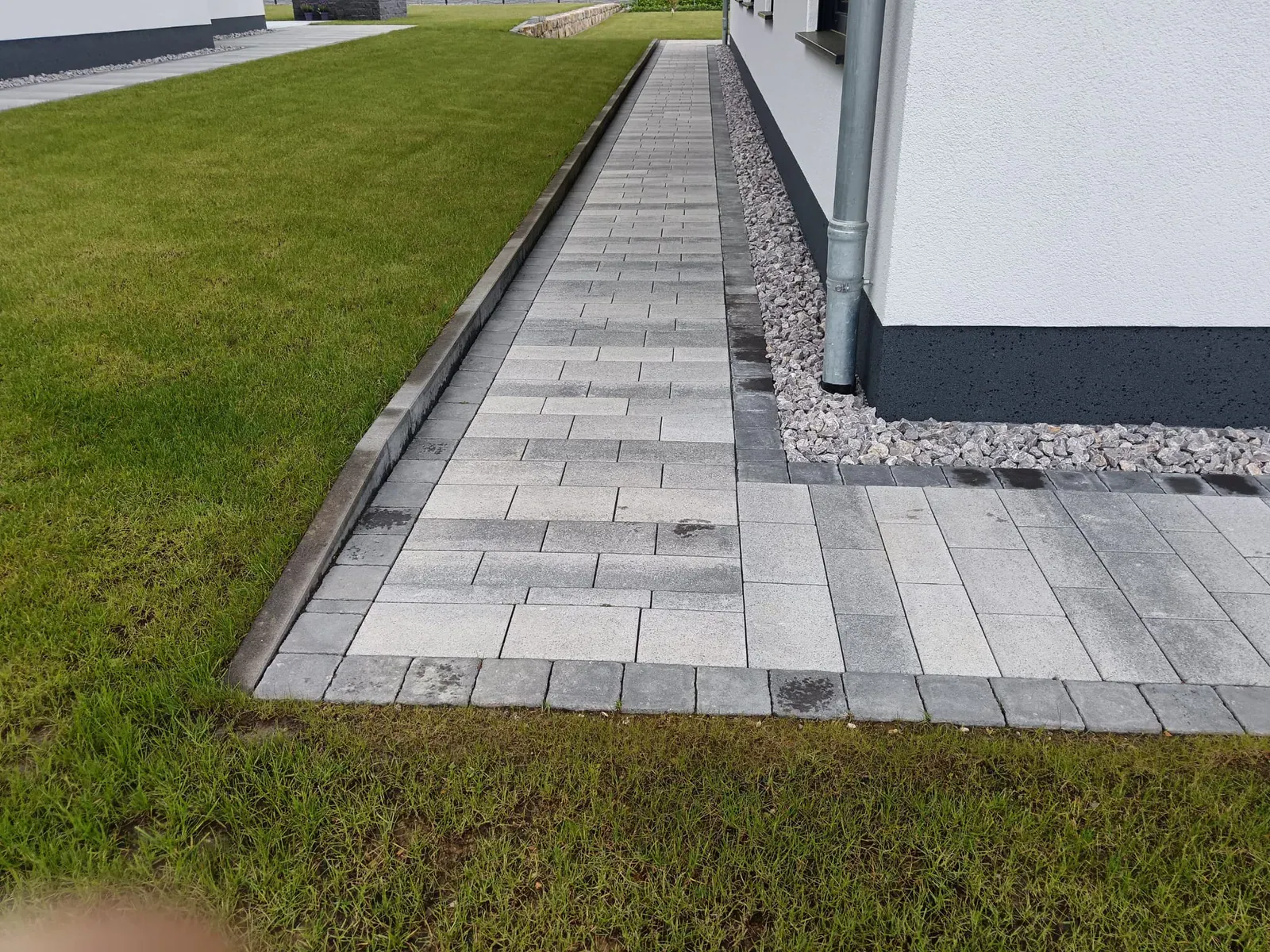 Gerader Seitenweg aus grauem Pflaster zwischen Hausfassade und gepflegter Rasenfläche.