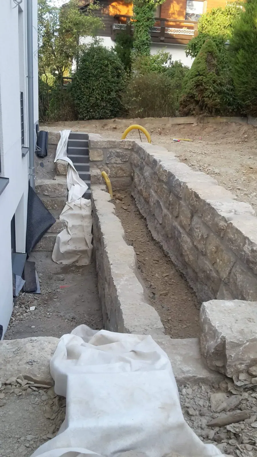 Dokumentation einer langen Natursteinmauer während der Ausführung mit vorbereitetem Graben und Hinterfüllung.