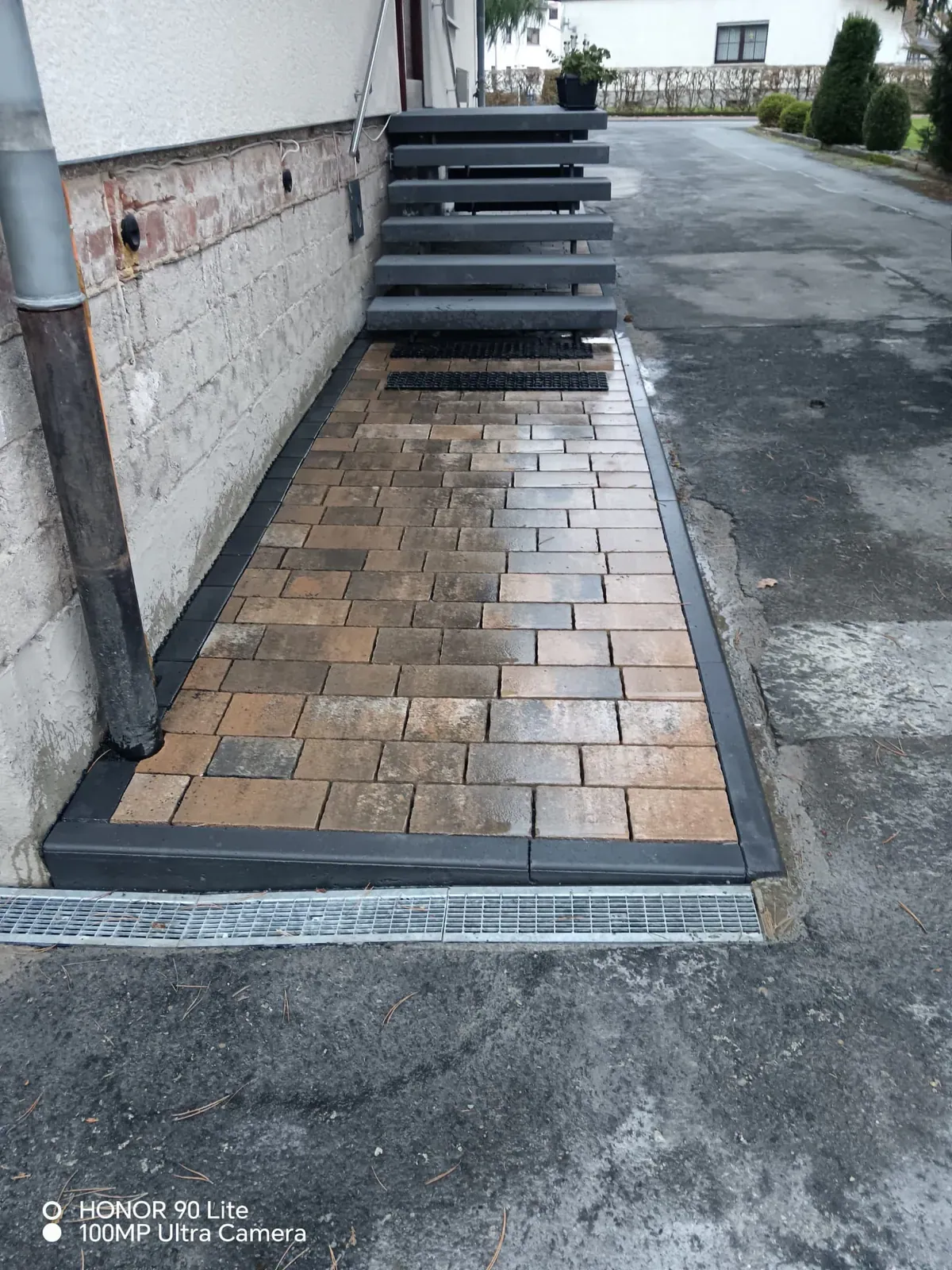 Schmaler Hausweg mit warmen Pflastertönen, dunkler Einfassung und angrenzender Asphaltfläche.