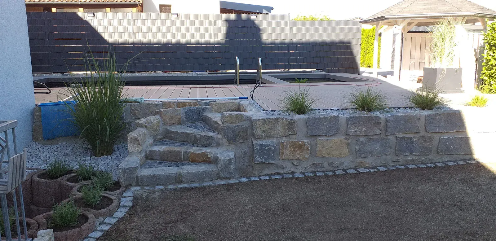 Fertige Poolumgebung mit rustikaler Natursteinmauer, Gräserbepflanzung und dunklem Sichtschutz.