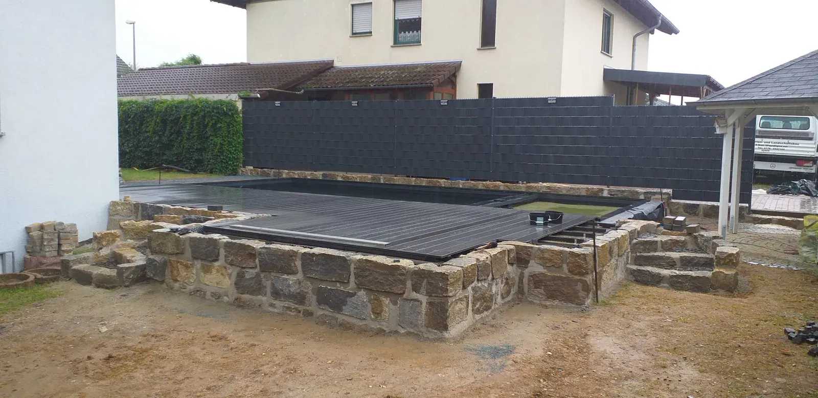 Dunkles Terrassendeck am Pool auf einer Einfassung aus Naturstein, ergänzt durch einen modernen Sichtschutz.