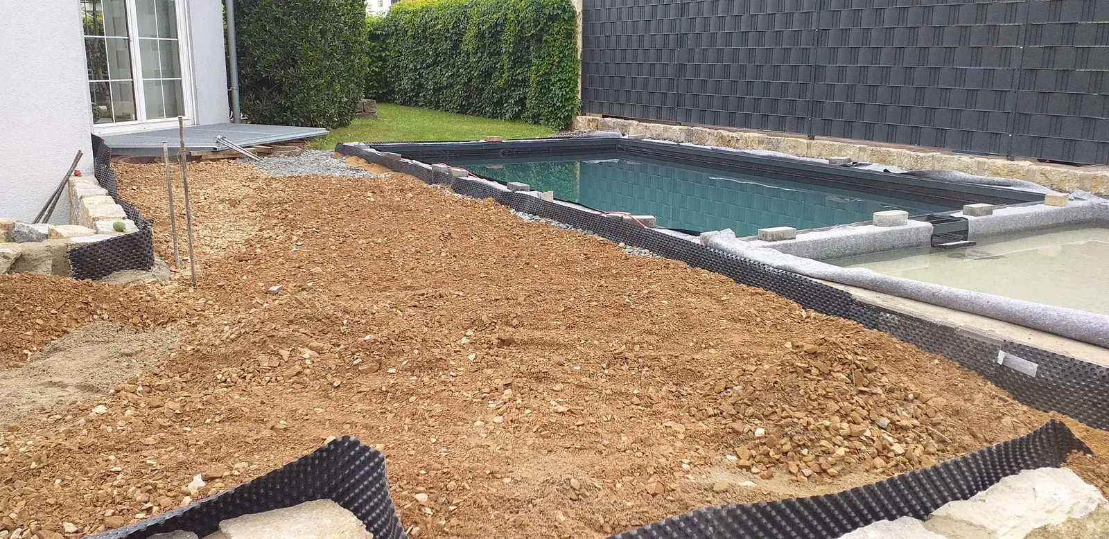 Außenpool mit Natursteinrand, angrenzendem Terrassenbereich und vorbereitetem Gartenboden.
