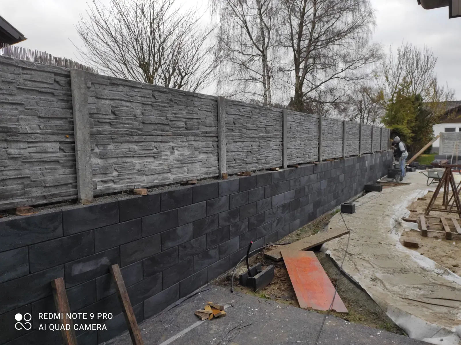 Lange Grundstückseinfriedung mit dunklem Sichtschutz, gemauertem Sockel und vorbereitetem Unterbau.