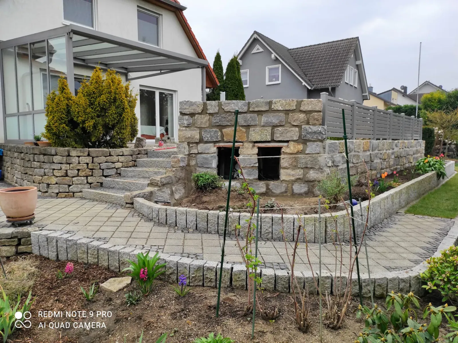 Abwechslungsreicher Vorgarten mit Natursteinabfangung, Blumenbeeten und gepflasterten Wegen.